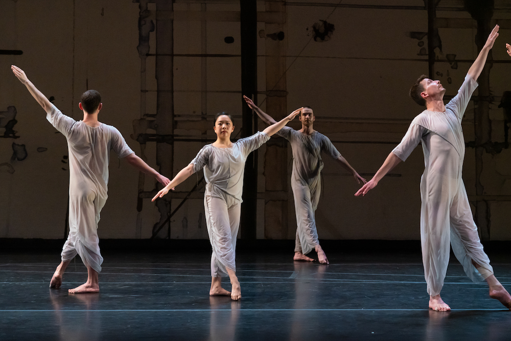 four dancers  stand on a stage wearing sheer blue-grey jump suits and pointing one foot forward, .they look directly into the audience with their arms spread in diagonal line from fingertip to finger tip...another dancer stands facing a side corner. we see him in profile standing similarly to the other three, his gaze follows the path if his higher arm, his lower arm behind him finishes the diagonal line 